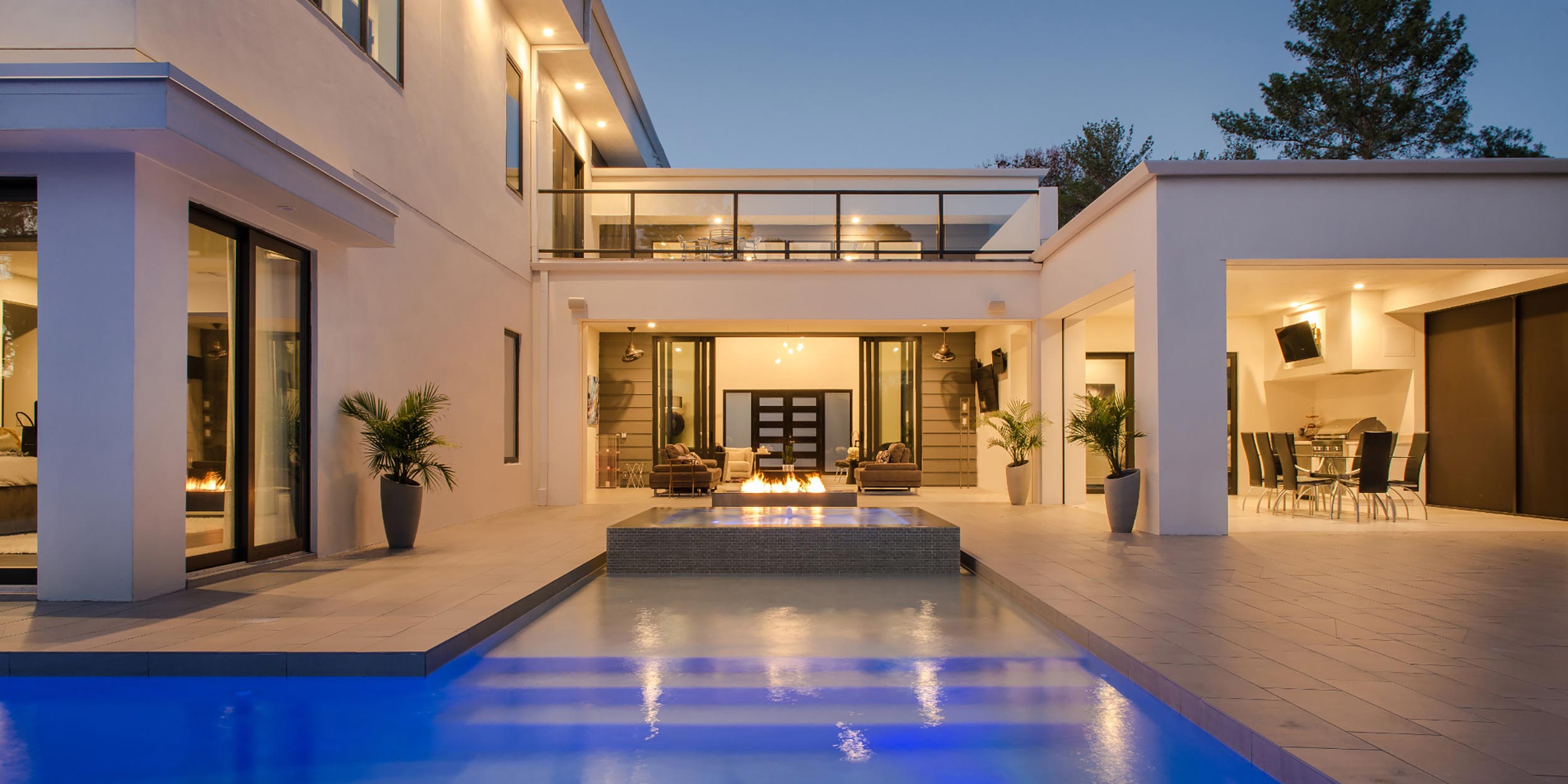 An outdoor modern house with an illuminated swimming pool and fire pit in the evening.