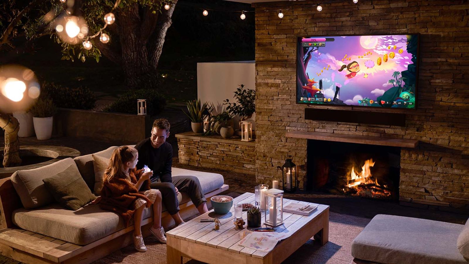 A father and daughter sitting by an outdoor fireplace under string lights, with a TV showing a vibrant video game.