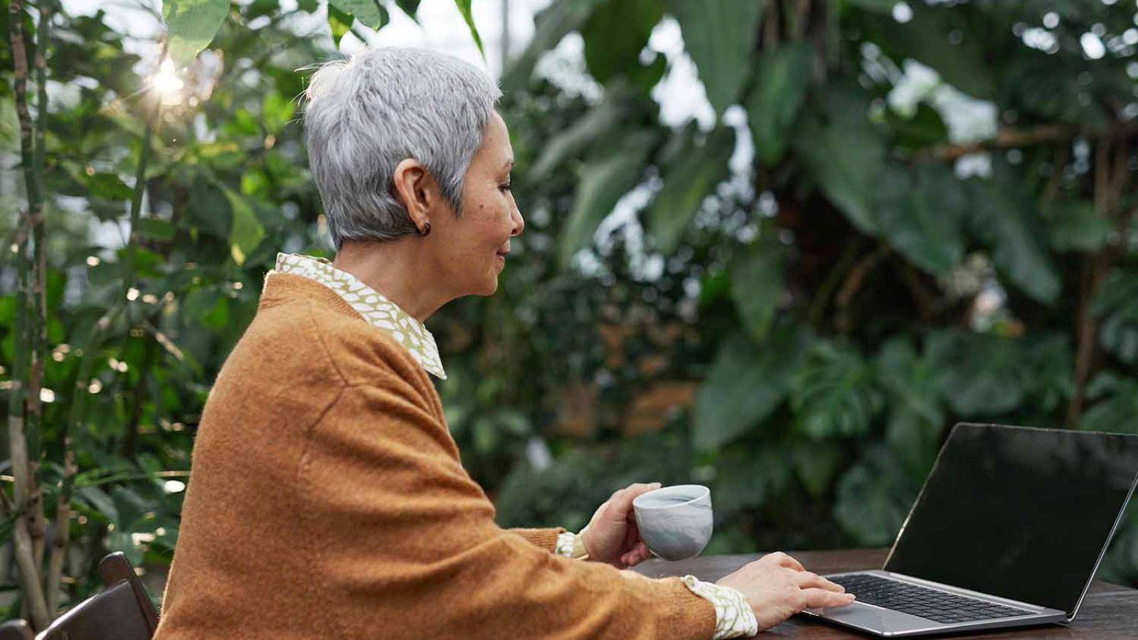 An older woman working on a laptop in a lush garden setting, holding a coffee mug and enjoying the greenery around her.